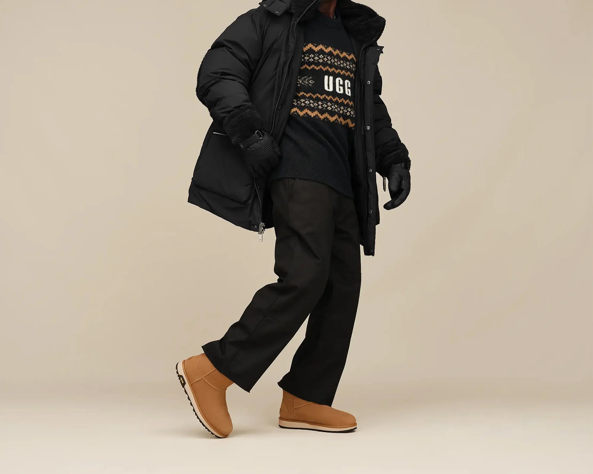 Person wearing tan suede ankle boots, black pants, black patterned sweater with "UGG" text, black puffer jacket, and black gloves.