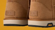 Close-up of tan suede UGG boots with white and black Vibram soles against a mustard background.