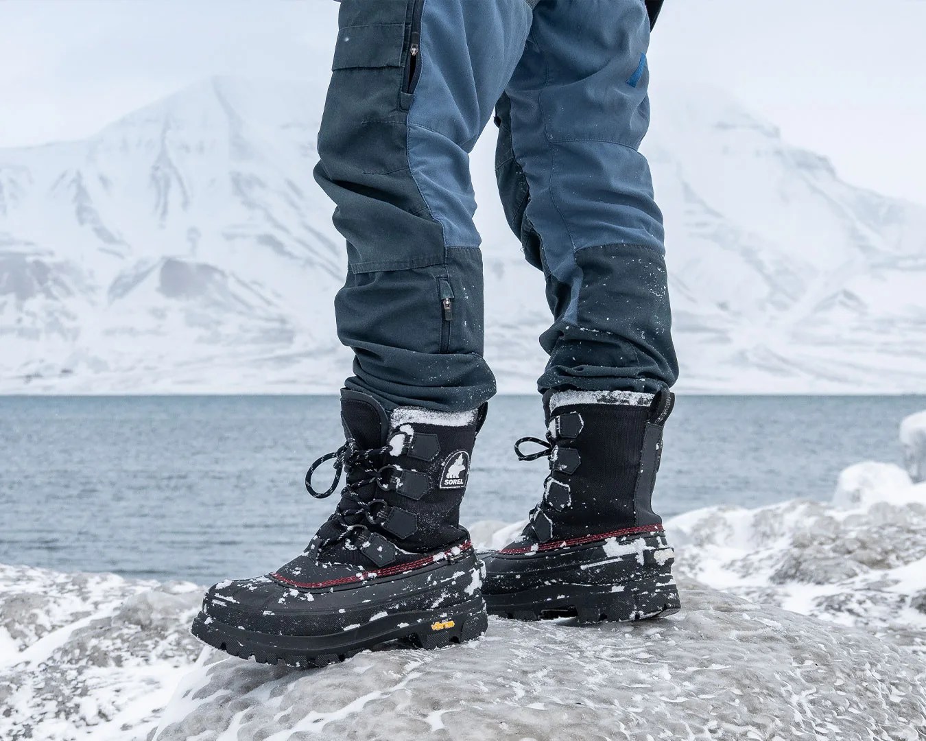 Black and red insulated winter boots with snow on them worn with blue snow pants on icy rocks by a lake.