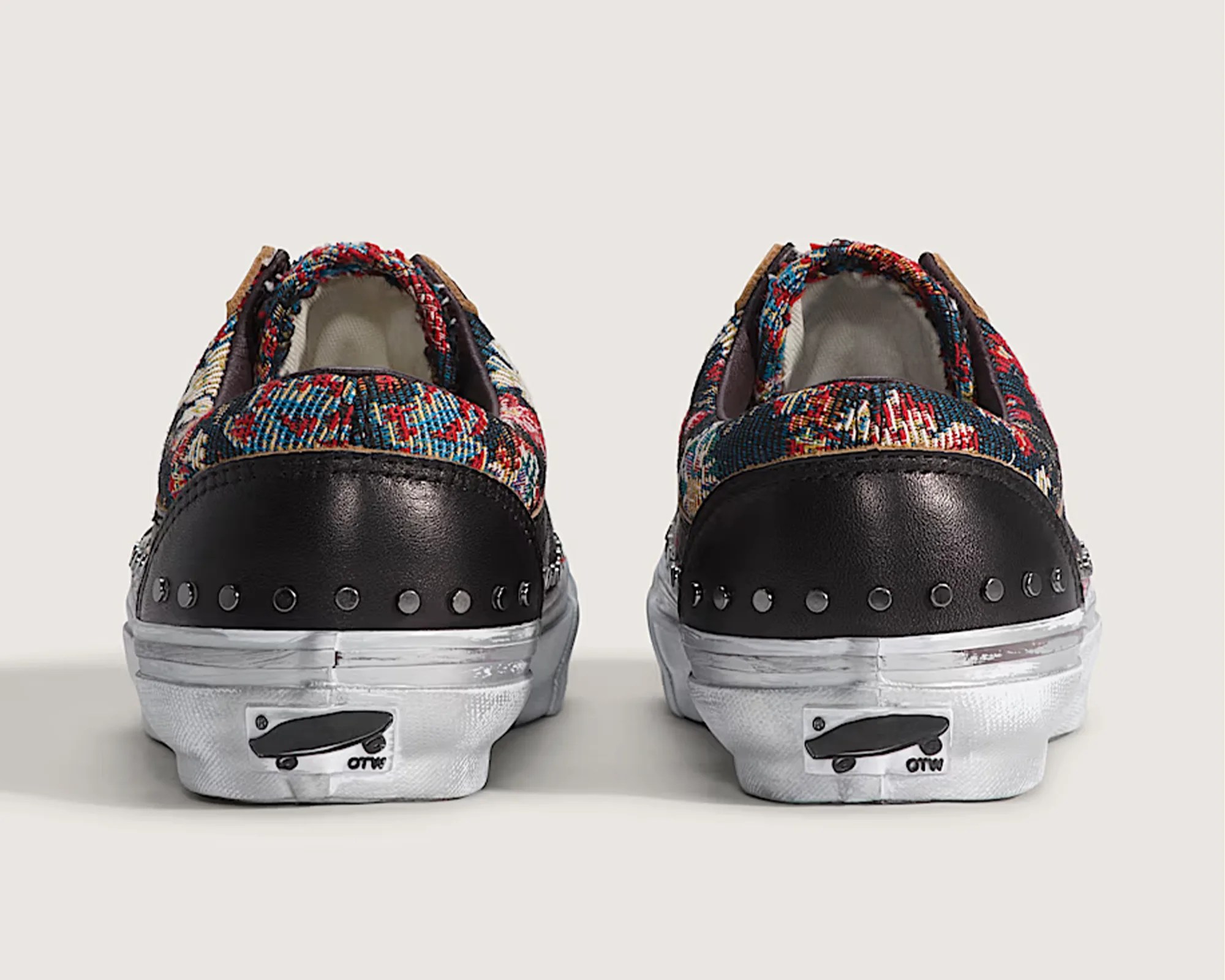 Pair of low-top sneakers with multicolor woven fabric, black leather heel panels with metal studs, and white rubber soles viewed from the back.