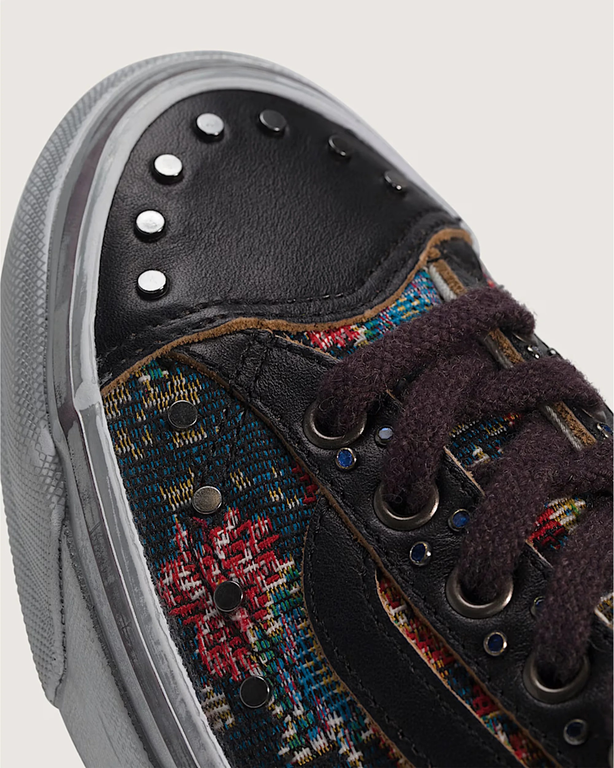 Close-up of a sneaker toe with black leather, multicolored woven fabric, silver studs, and dark purple laces.