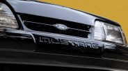 Close-up of a black Ford Mustang front grille with headlights and a yellow background.