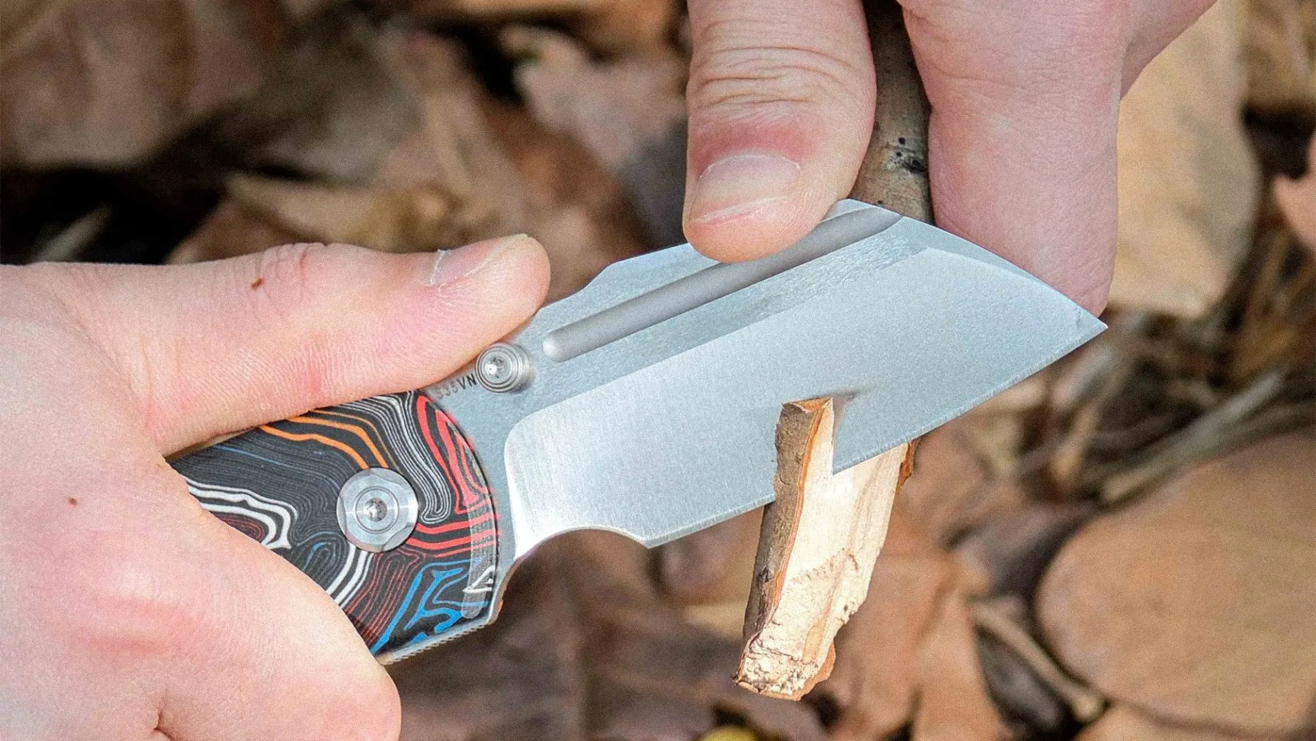 Small folding knife with a multicolored handle cutting a small piece of wood.
