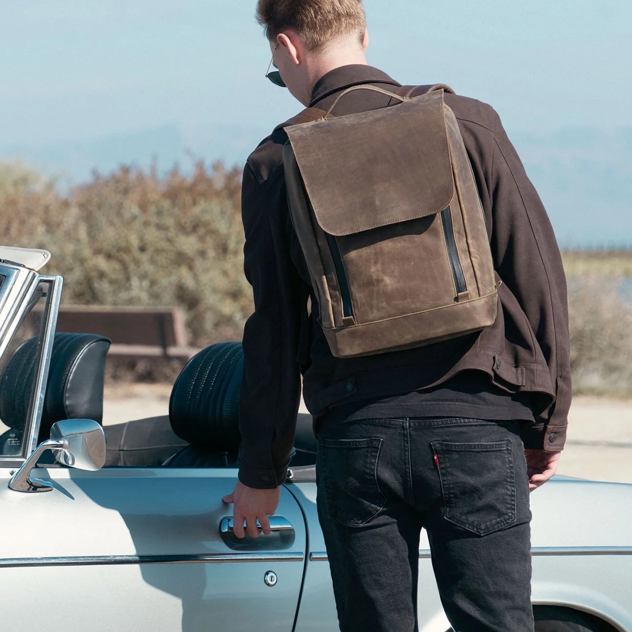 Brown leather backpack with flap and vertical zippers worn by a person opening a car door.