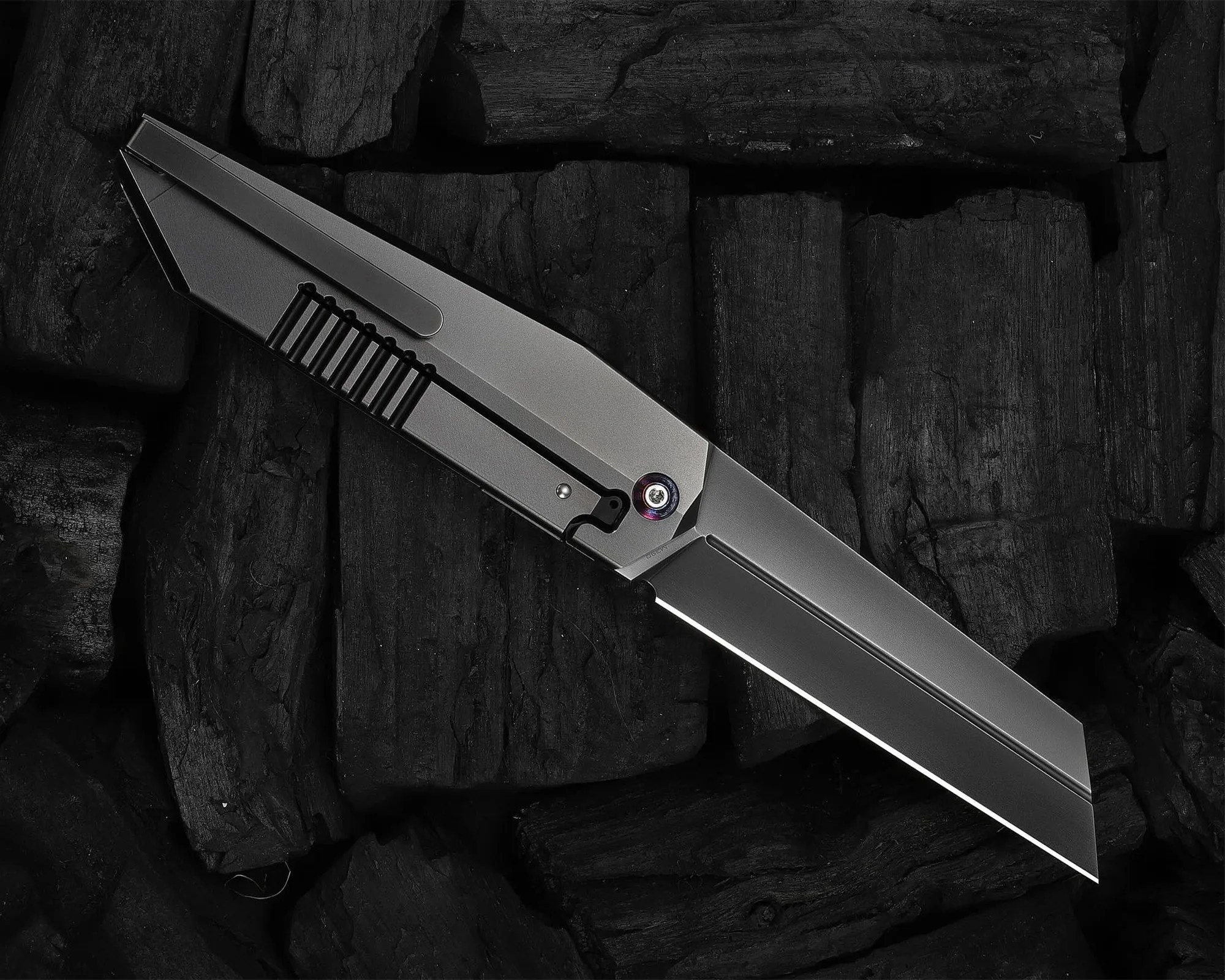 Sleek metallic folding knife with a straight-edged blade resting on dark charcoal pieces.