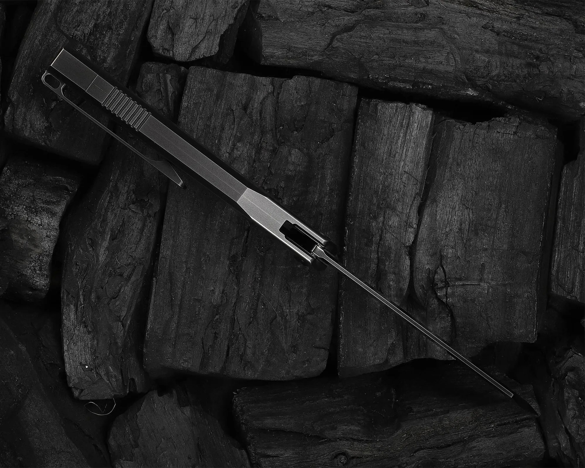 Slim metallic folding knife with a long, thin blade resting on black charcoal pieces.