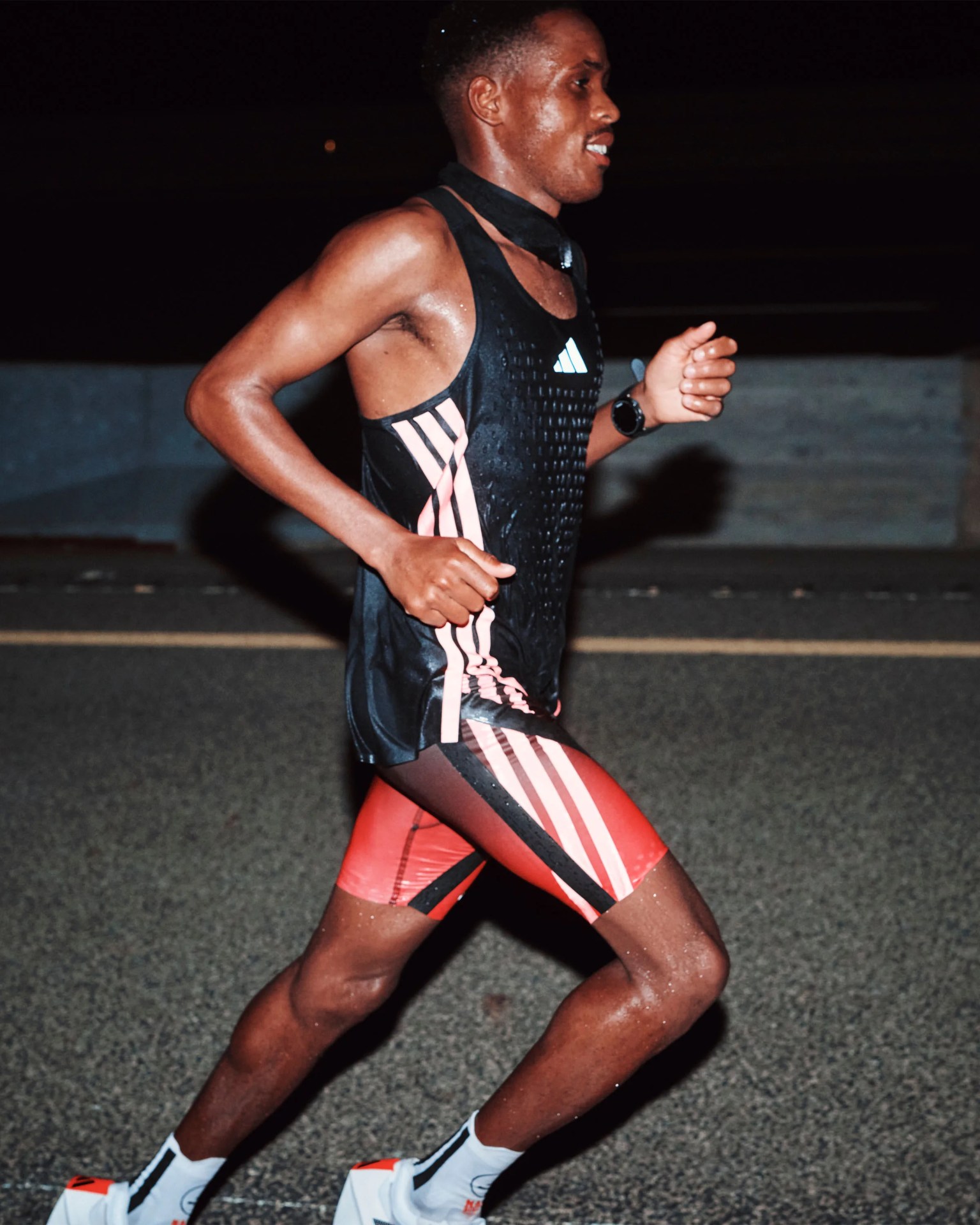 Runner wearing a black and red Adidas athletic outfit and white running shoes on a road at night.