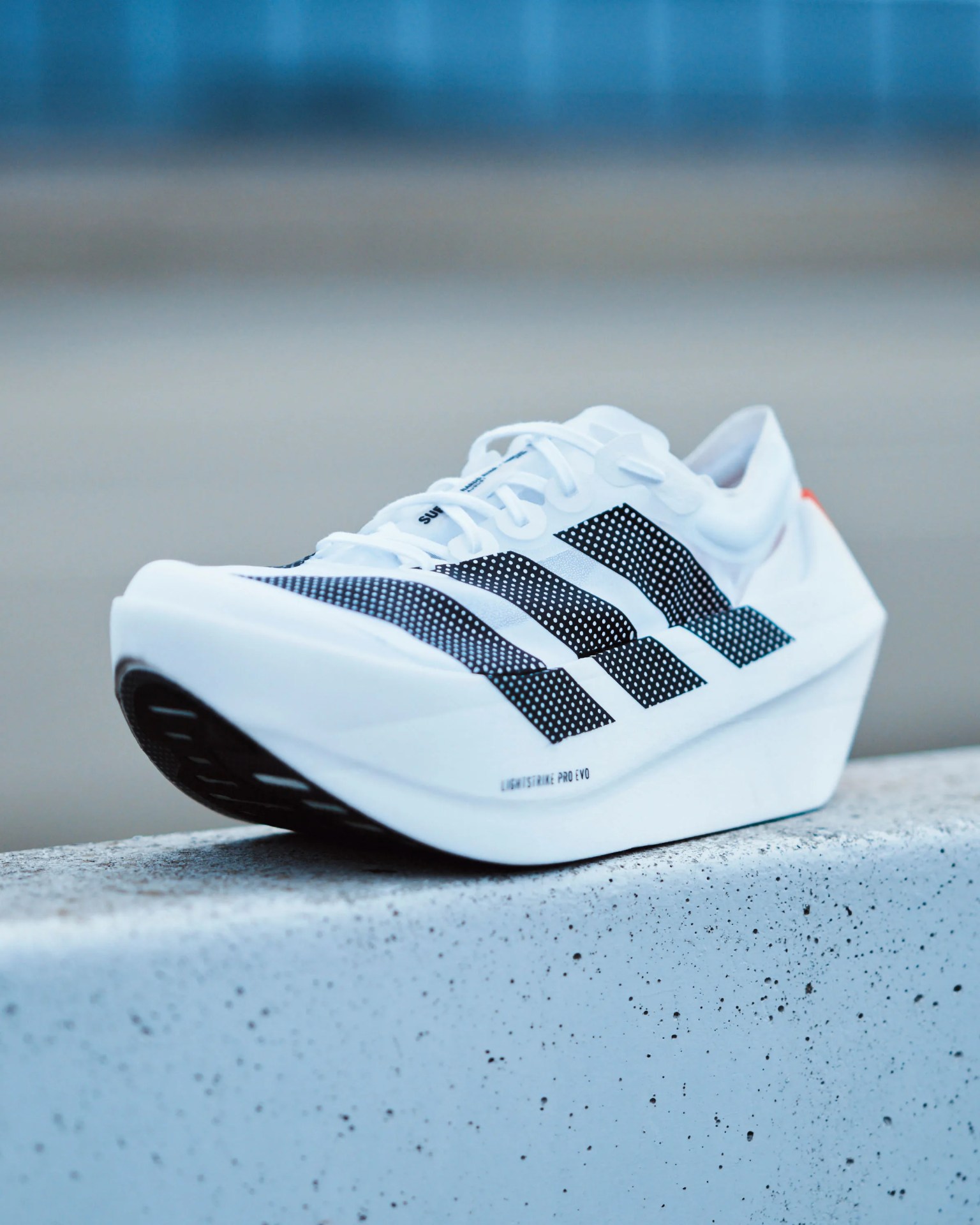 White Adidas running shoe with black three stripes and thick sole resting on a speckled concrete surface.
