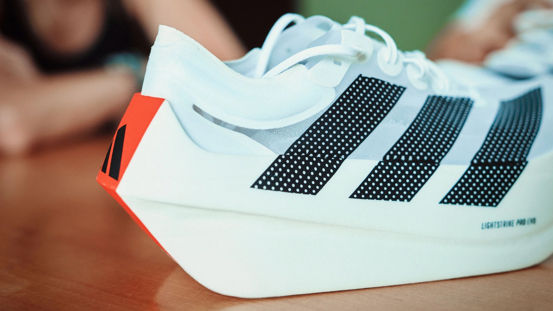 White Adidas running shoe with black dotted stripes and orange heel detail on a wooden surface.