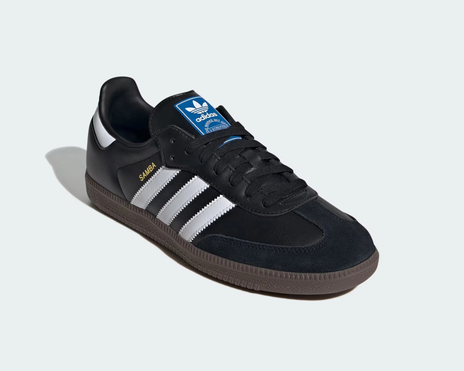 Black Adidas Samba sneaker with white stripes, gold "SAMBA" text, blue logo patch on tongue, and brown sole.