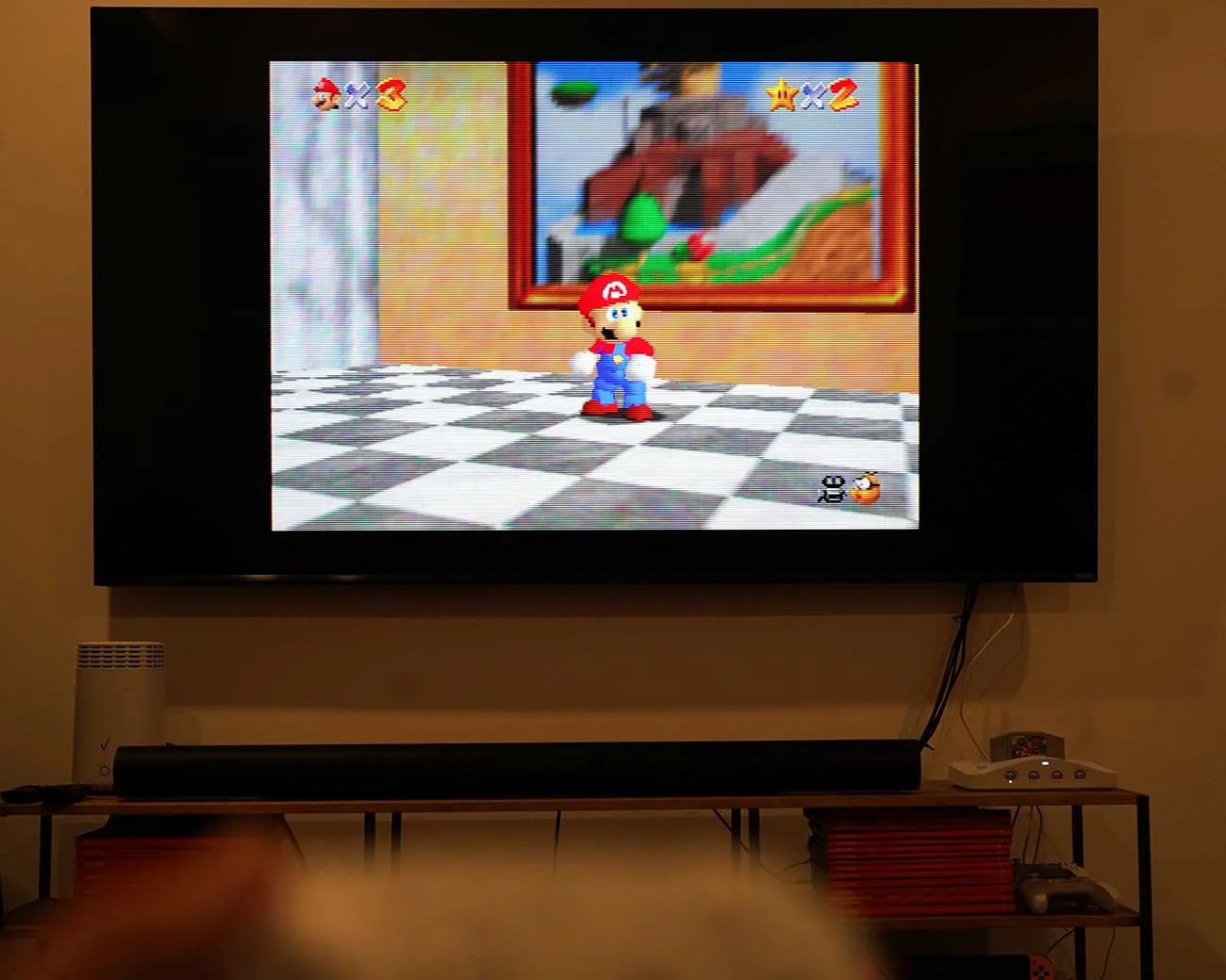 Flat-screen TV mounted on a wall displaying the video game Super Mario 64 with a Nintendo 64 console and soundbar below.