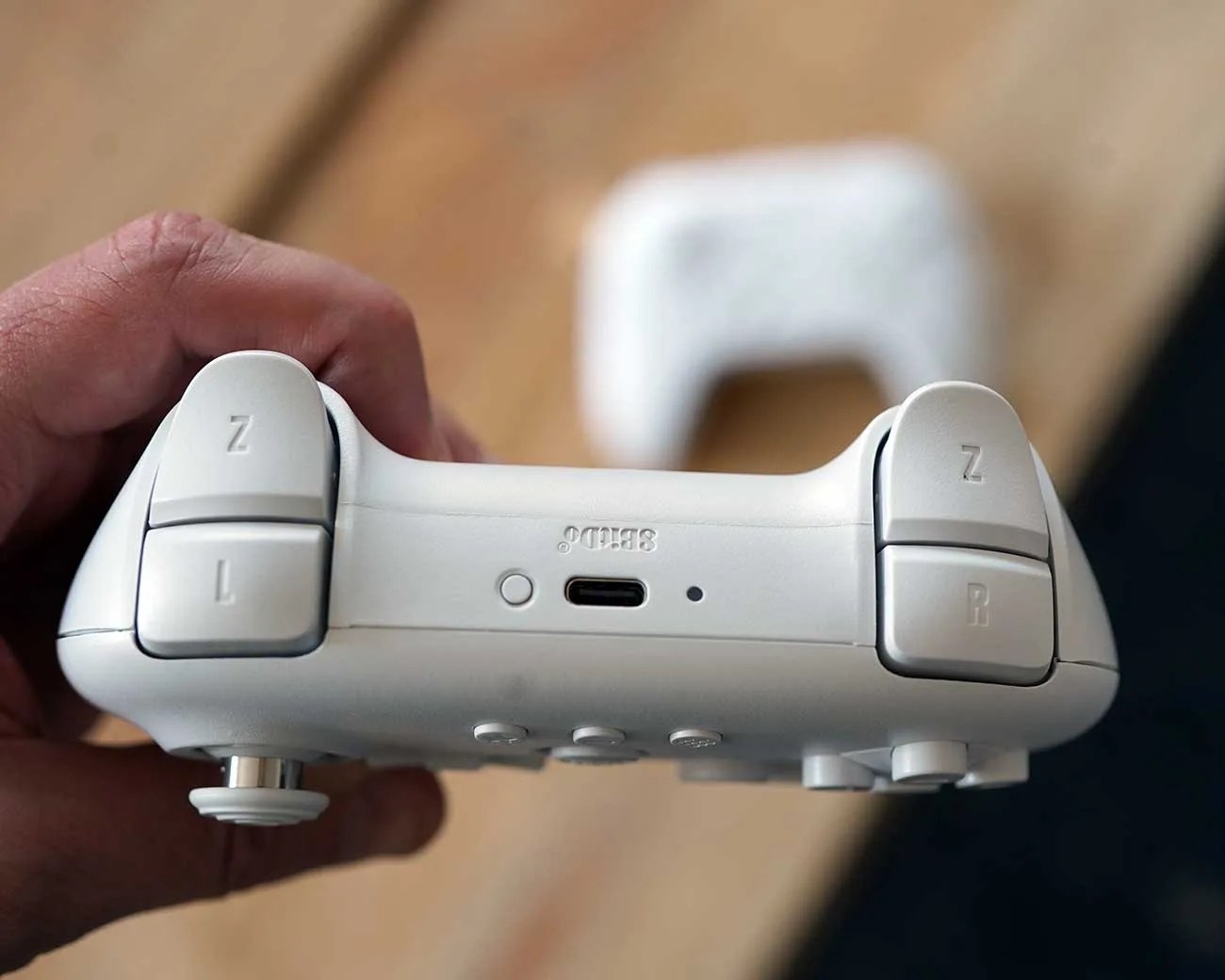 White gaming controller held in a hand showing top side with L and R buttons and a USB-C charging port.
