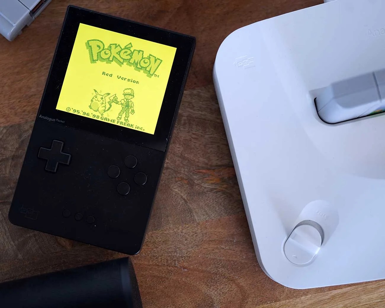 Black Analogue Pocket handheld console displaying Pokémon Red Version on screen, next to a white device on a wooden surface.