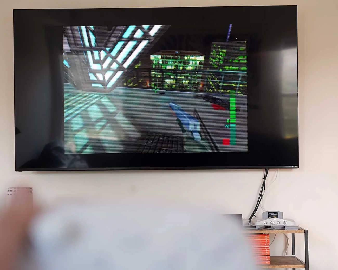 Flat-screen TV mounted on a wall displaying a first-person shooter video game with a handgun and cityscape, with a Nintendo 64 console on a shelf below.