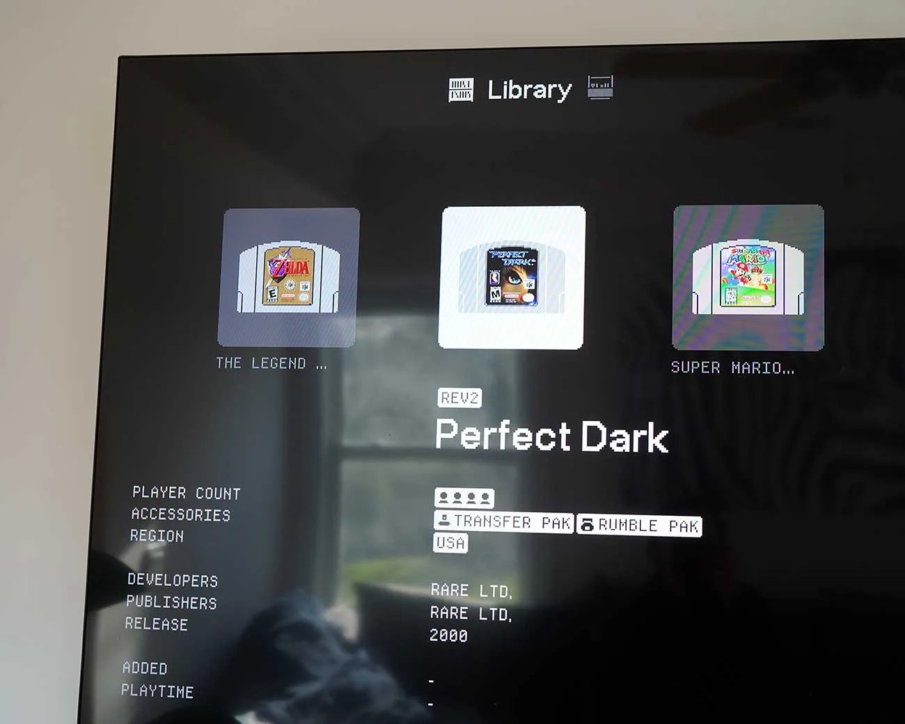 TV screen showing a game library menu with Nintendo 64 game cartridges for The Legend of Zelda, Perfect Dark, and Super Mario 64.