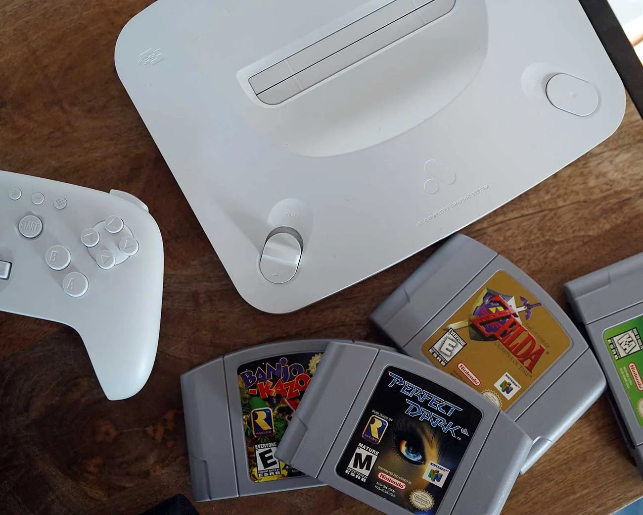 White retro gaming console with a matching white controller and three Nintendo 64 game cartridges on a wooden surface.