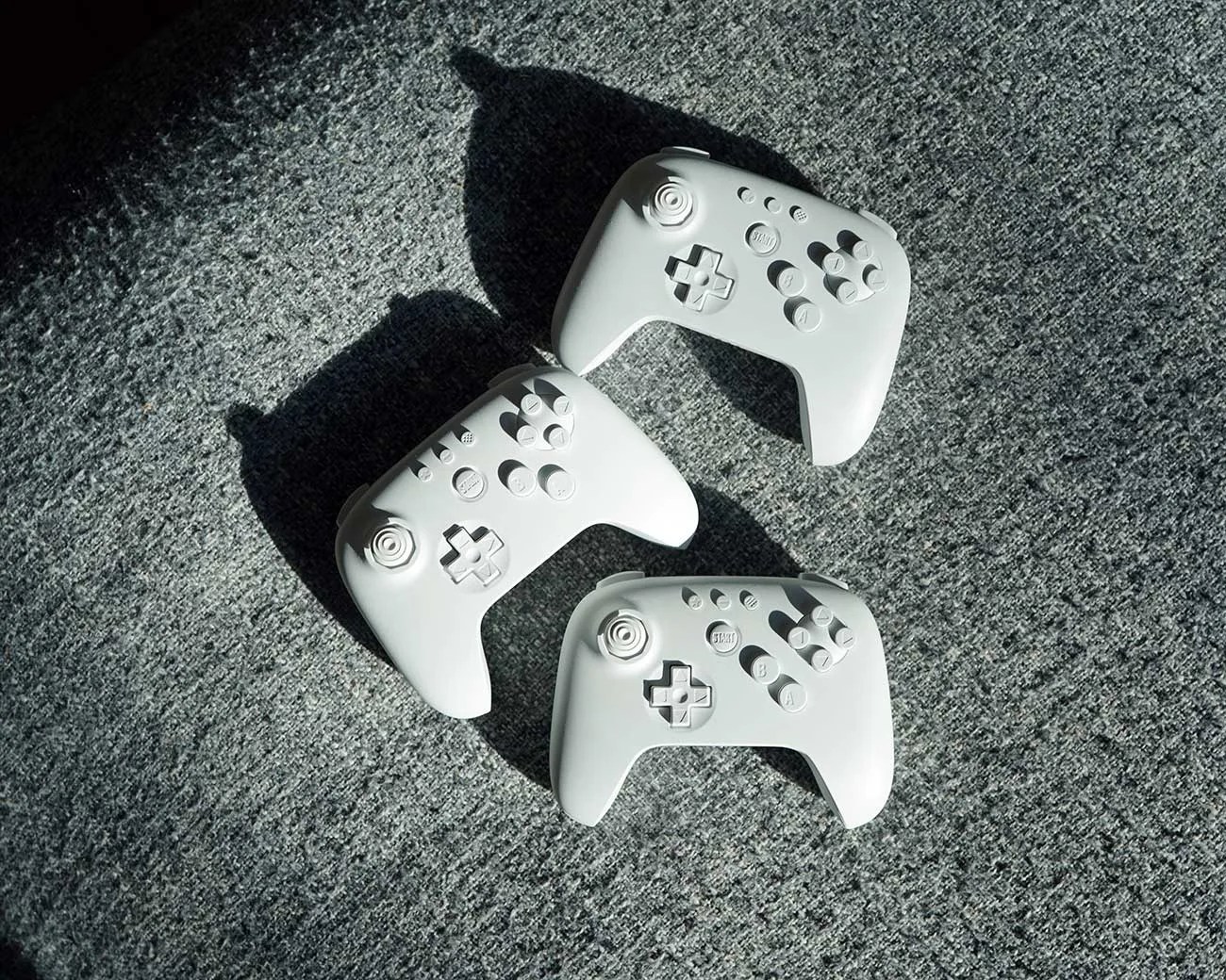 Three white wireless gaming controllers with buttons and joysticks on a gray textured surface.