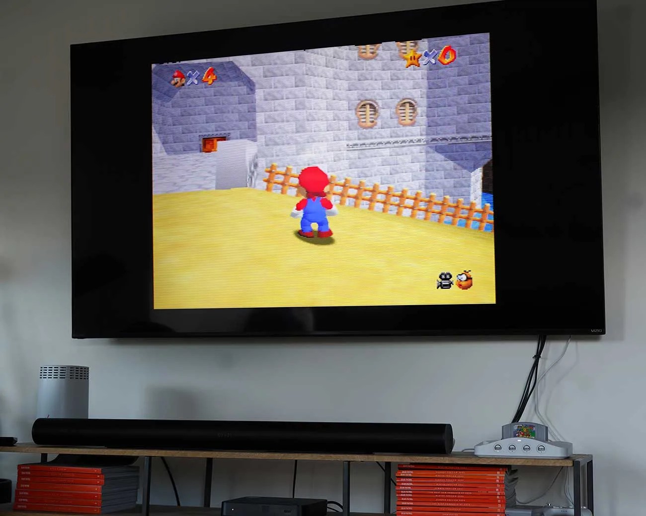 Flat-screen TV mounted on a wall displaying Super Mario 64 gameplay with a Nintendo 64 console on a wooden shelf below.