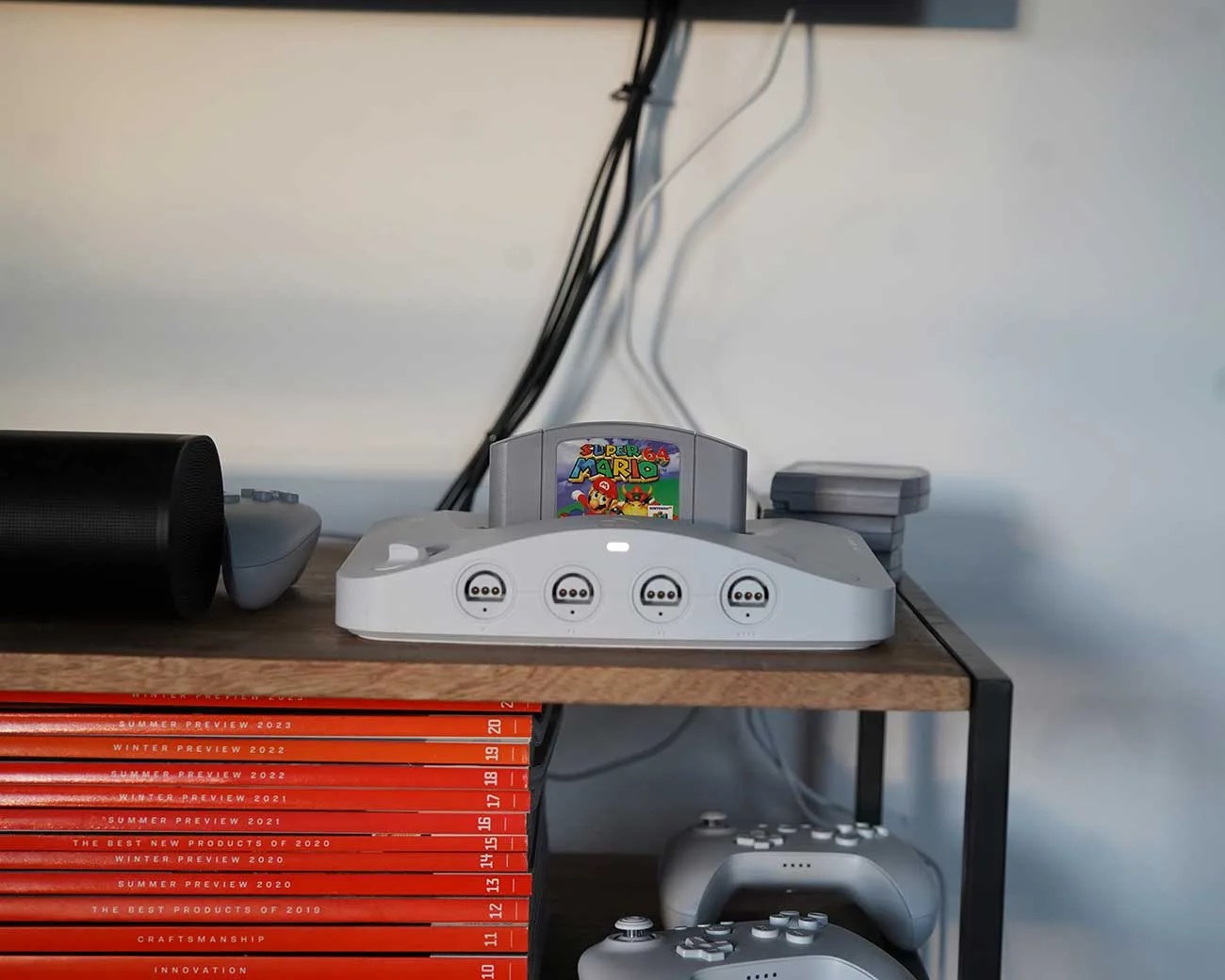 Gray Nintendo 64 console with Super Mario 64 cartridge on a wooden shelf with stacked game cartridges and controllers.