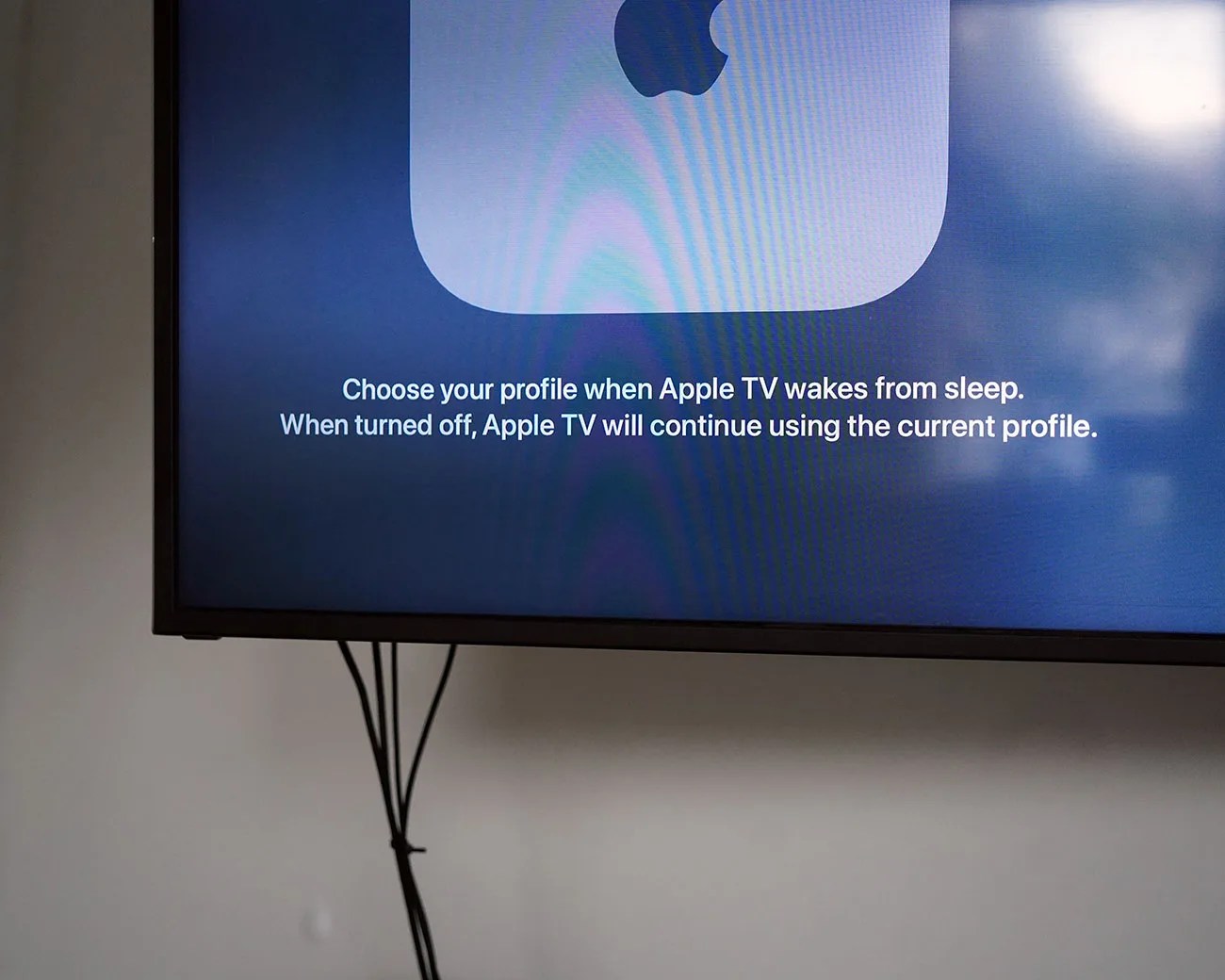 Television screen displaying Apple TV message about choosing a profile when waking from sleep, with visible cables below.