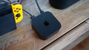 Black Apple TV device with cables connected, placed on a wooden surface next to a yellow Nintendo Switch controller.