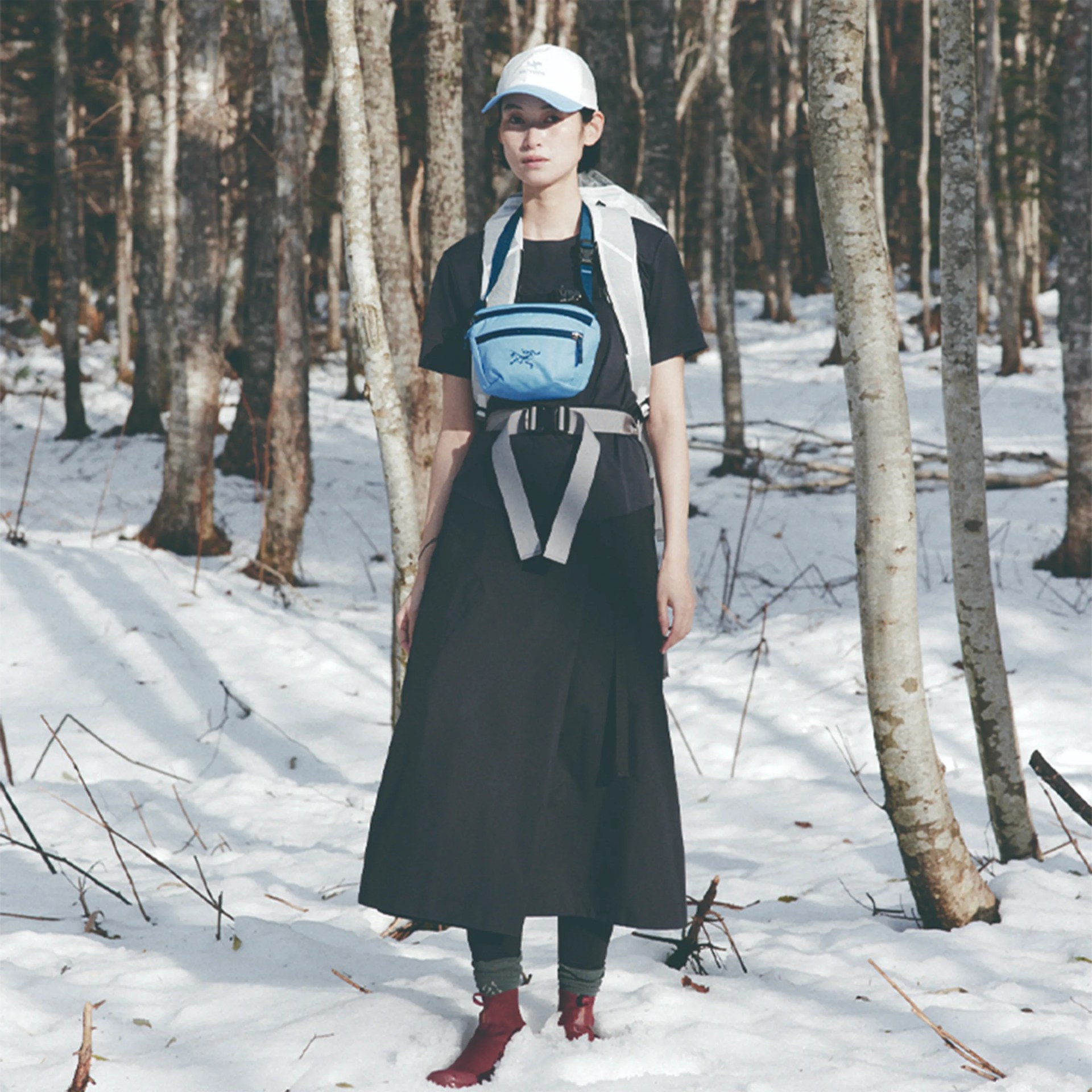 Person wearing a black dress, gray backpack, blue chest pouch, white cap, and red boots standing in a snowy forest.