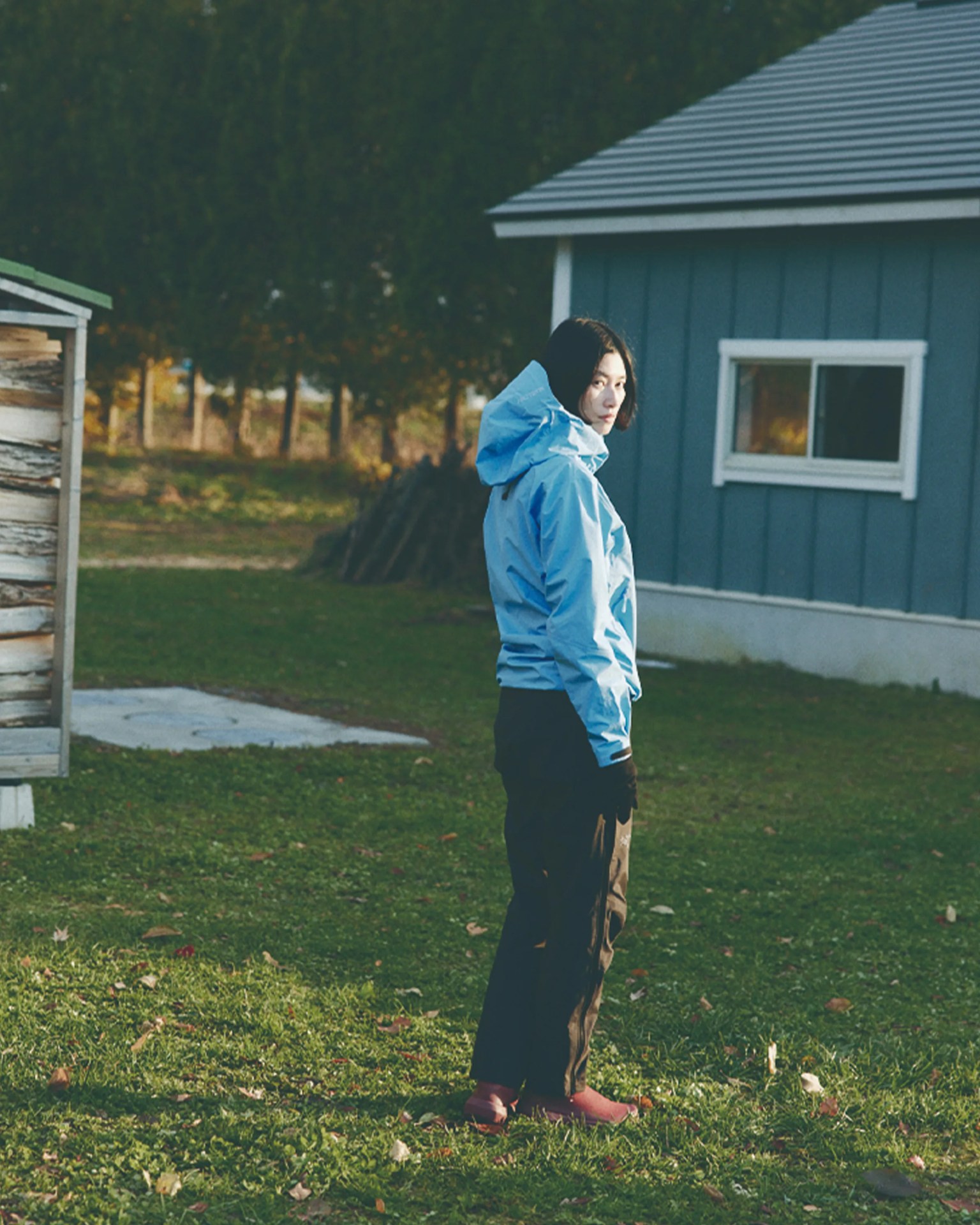 Person wearing a light blue hooded jacket, black pants, and red shoes standing on grass near a blue building.