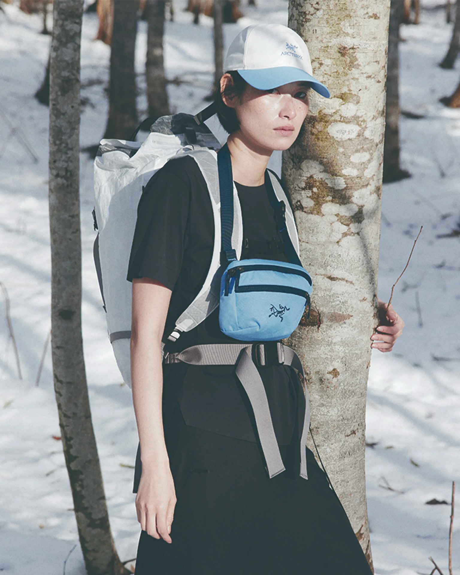 Person wearing a white and blue cap, black shirt, white backpack, and light blue chest bag standing in a snowy forest.