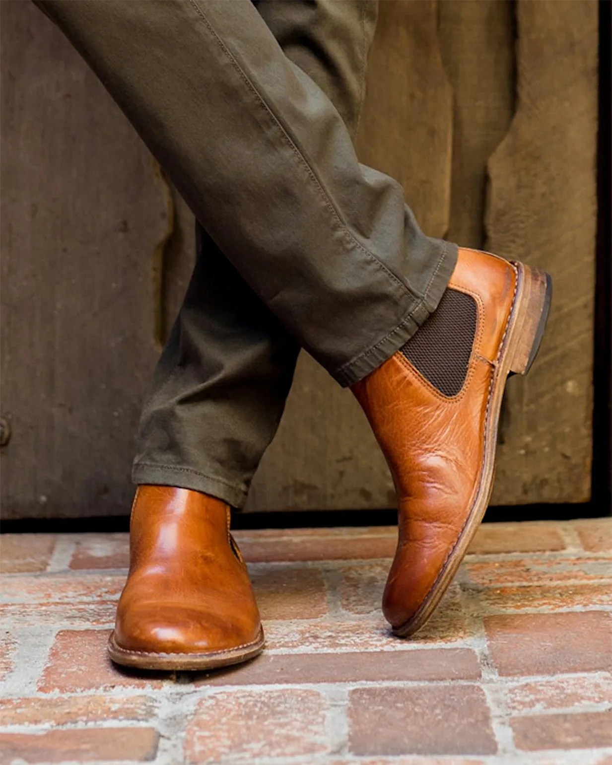 Pair of tan leather Chelsea boots with dark elastic side panels worn with olive pants on a brick floor.