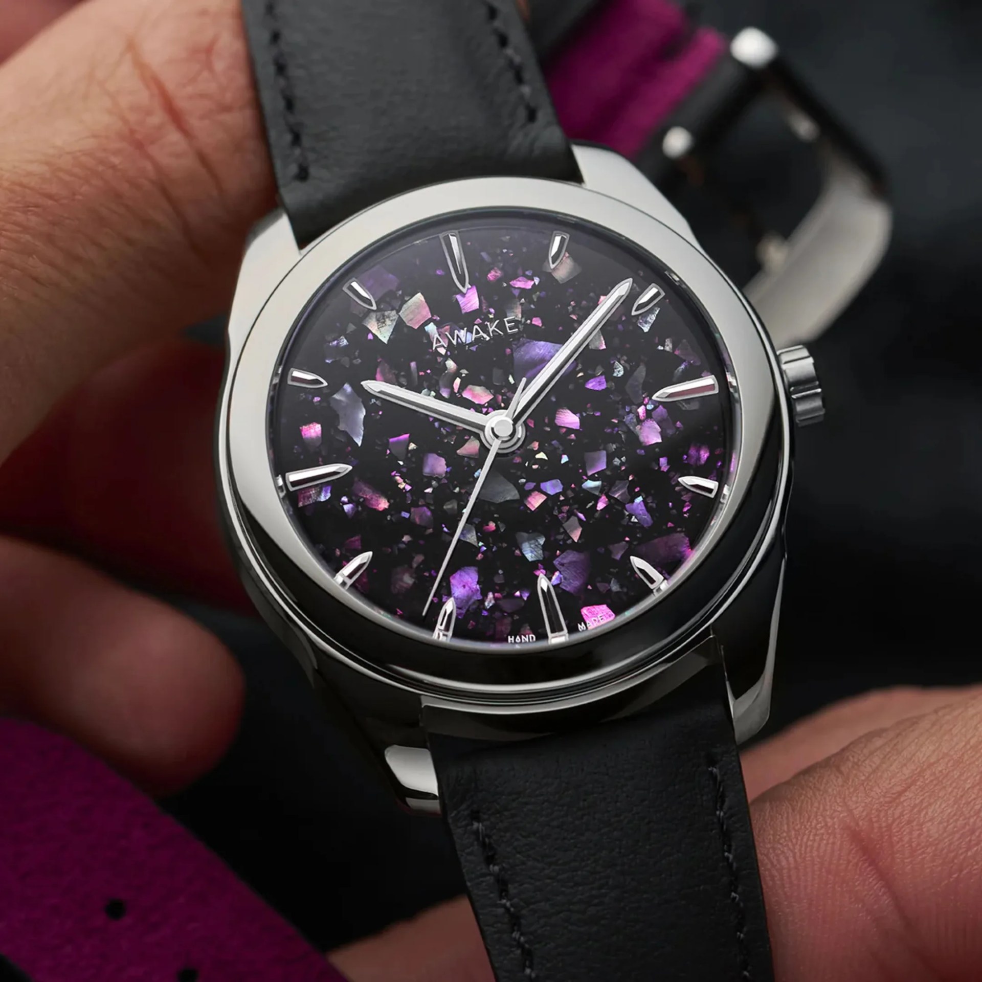 Close-up of a wristwatch with a black leather strap and a black dial featuring iridescent purple and pink fragments.