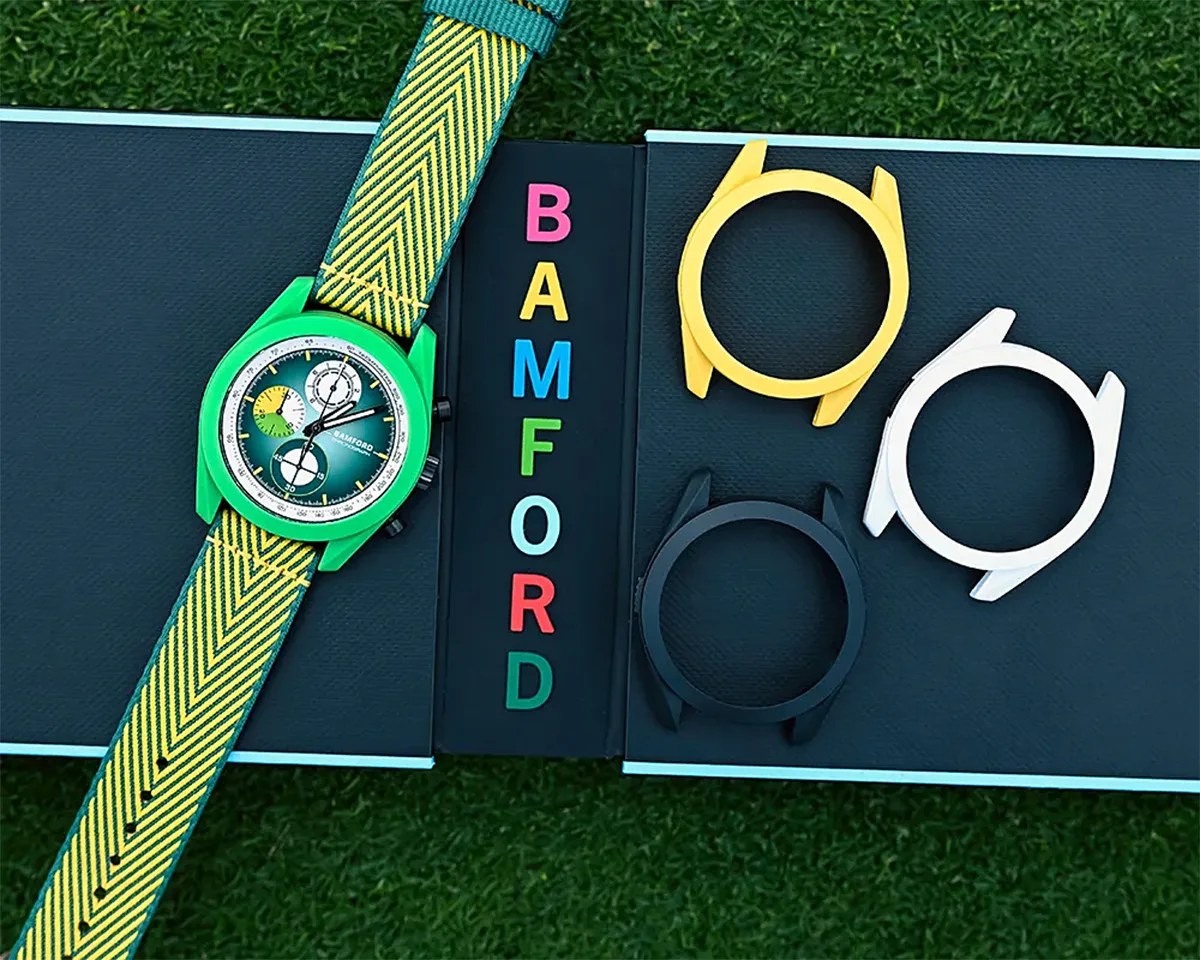 Green Bamford wristwatch with yellow and green chevron strap next to black box with colorful BAMFORD letters and three extra watch cases in yellow, black, and white.