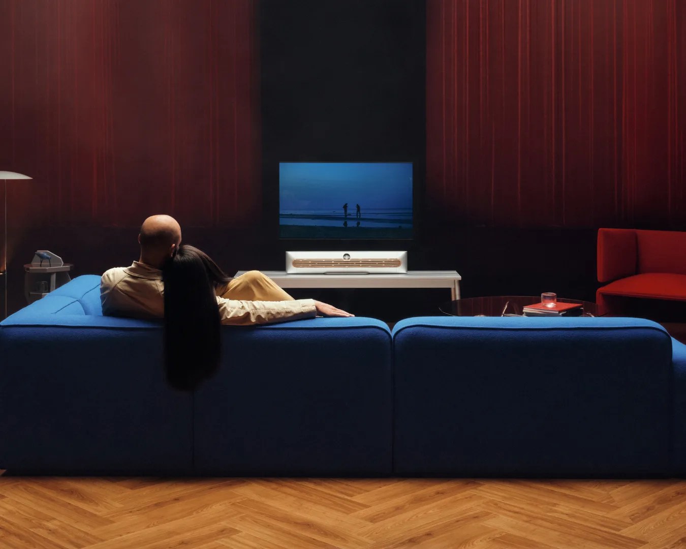 Couple sitting on a blue sofa watching a wall-mounted TV with a white soundbar on a white table in a dim room.