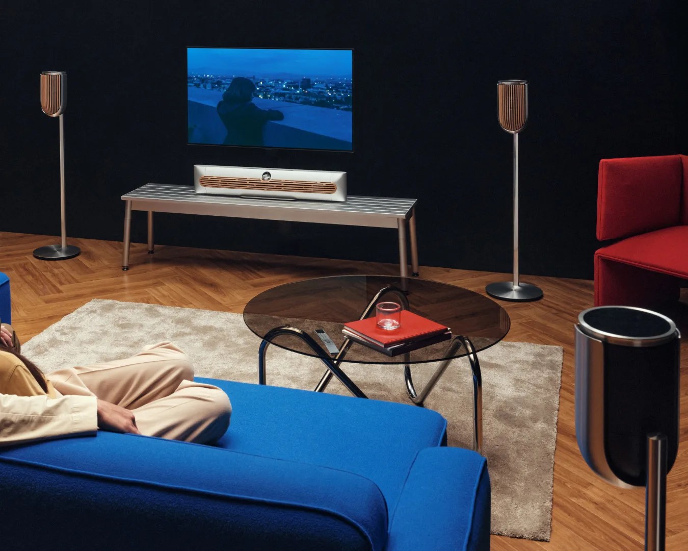 Modern living room with a blue sofa, glass coffee table, red armchair, and a TV with wooden-accented speakers.