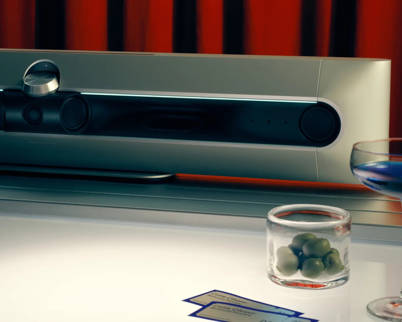 Silver and black soundbar with a volume knob on a table next to a glass bowl of green olives and a cocktail glass.