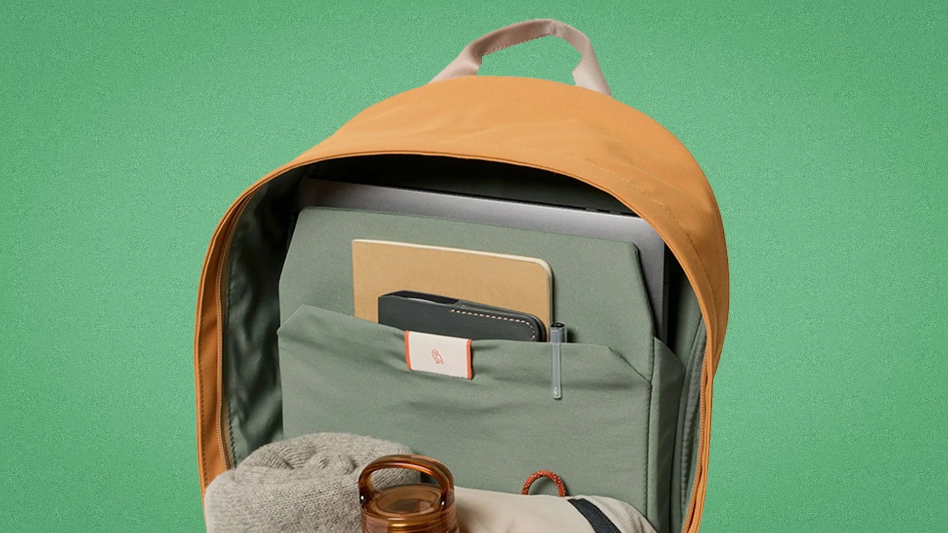 Orange backpack with green interior holding a laptop, notebook, wallet, pen, water bottle, and rolled sweater.