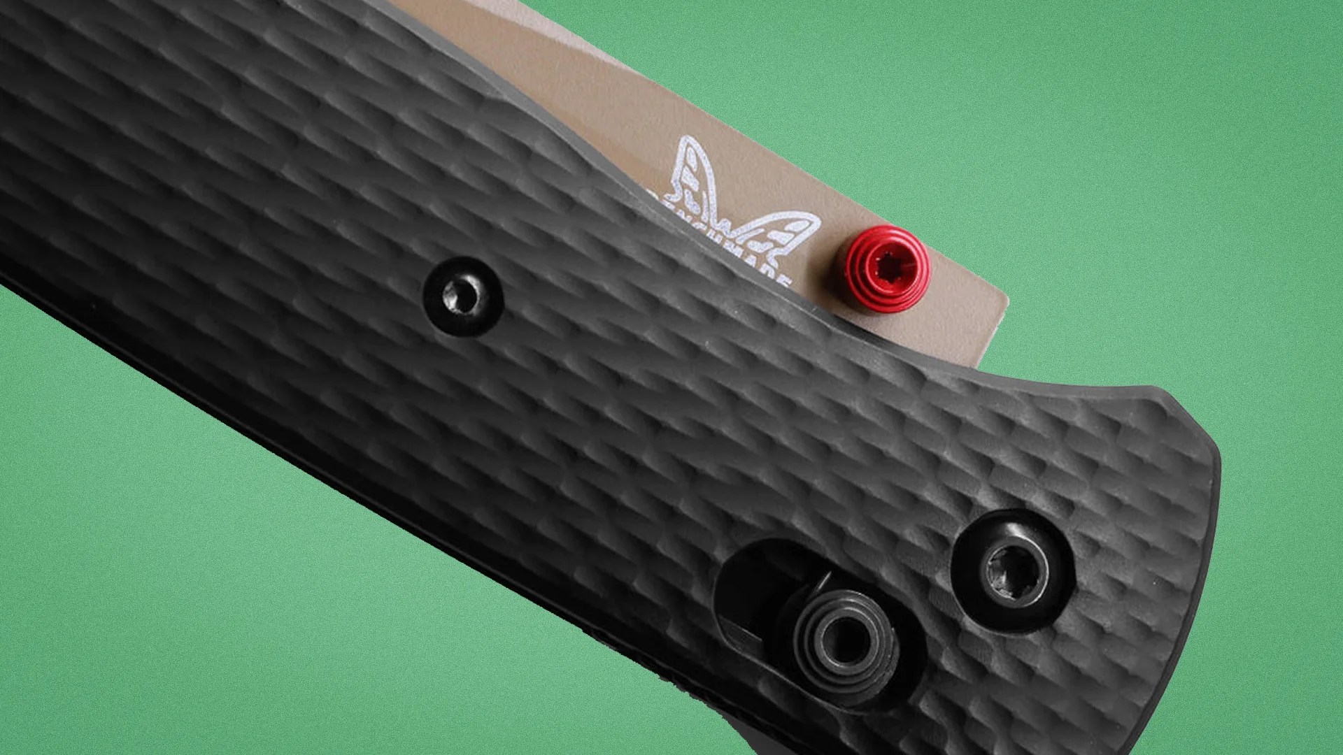 Close-up of a textured black knife handle with visible screws and a partially visible tan blade with a red thumb stud.