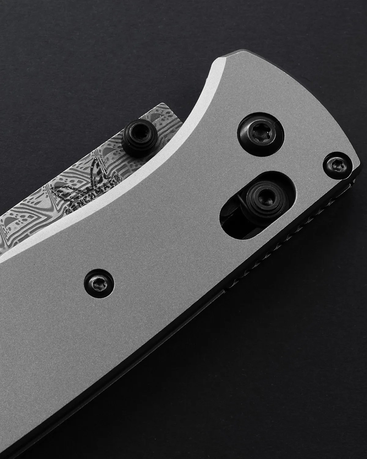 Close-up of a folding knife with a gray handle and a partially visible patterned blade on a black background.