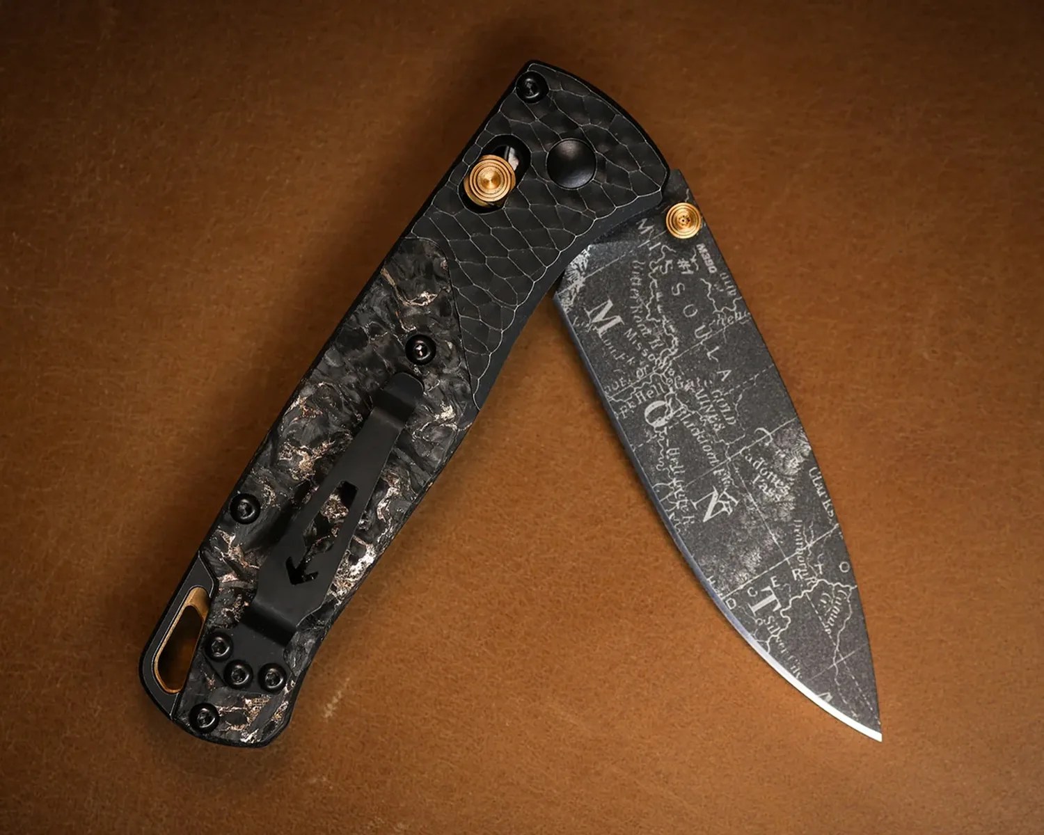 Folding knife with black textured handle and blade etched with a vintage map design on a brown surface.
