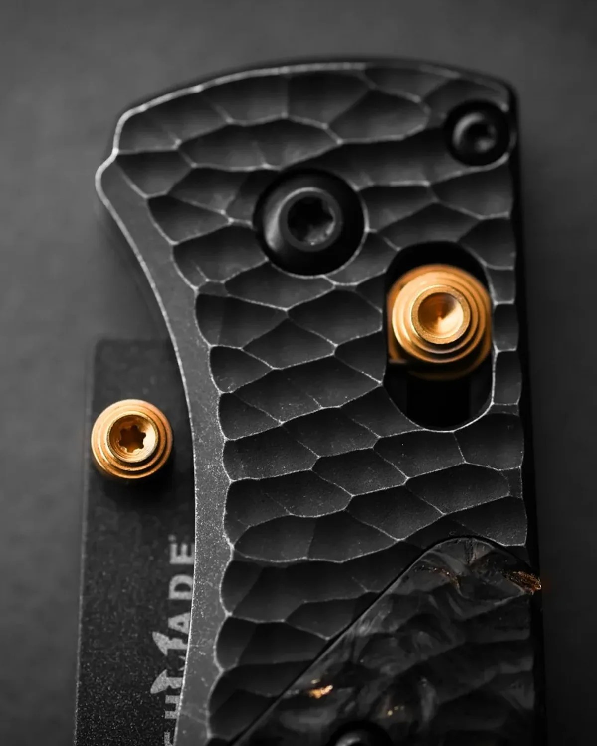Close-up of a textured black metal surface with two gold-colored screws and a partially visible logo on a dark background.