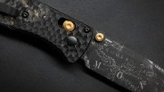 Close-up of a black folding knife with textured handle, gold pivot screws, and a blade etched with map-like markings.