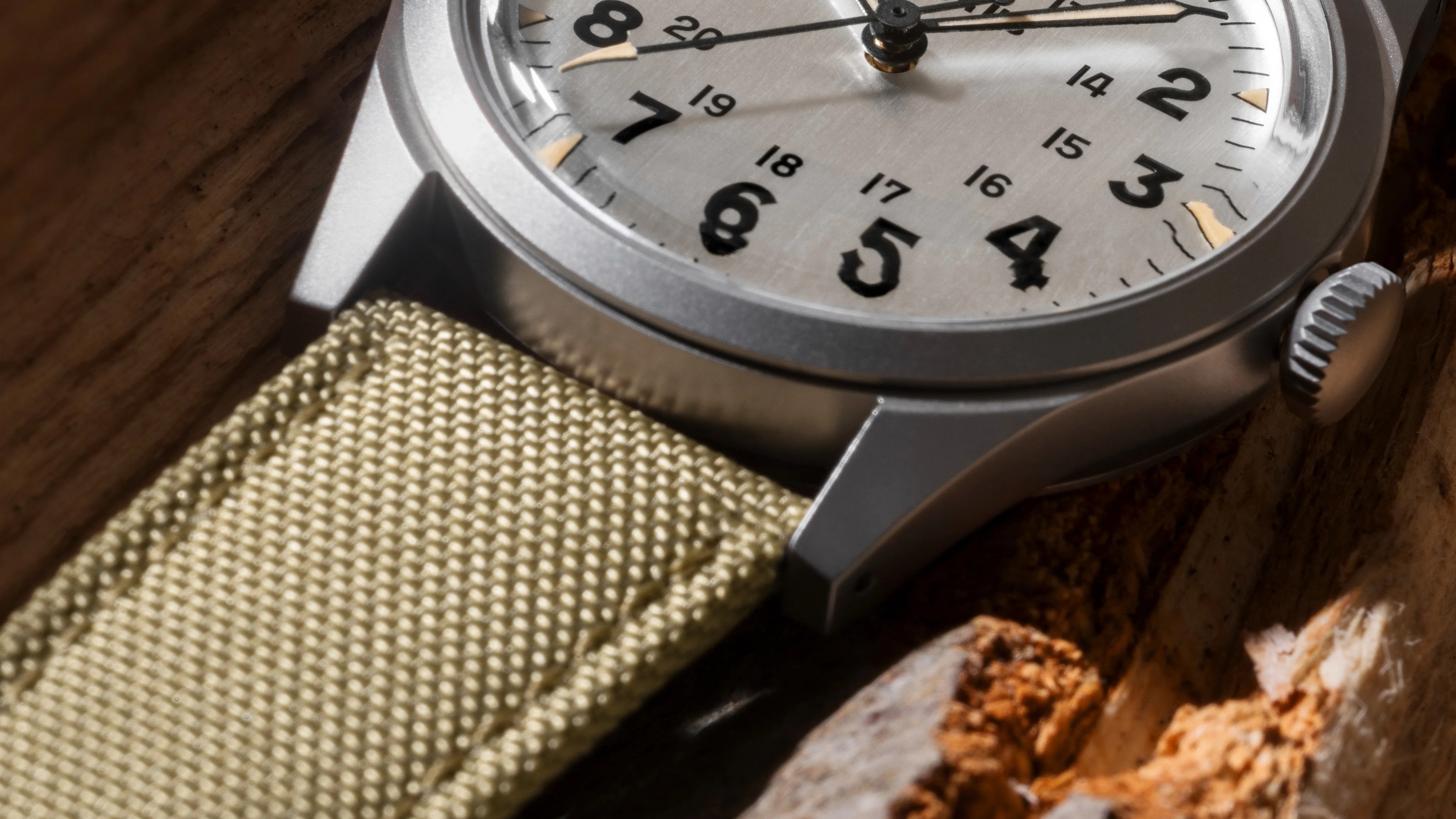 Close-up of a silver watch with black numbers and a beige woven fabric strap on a wooden surface.