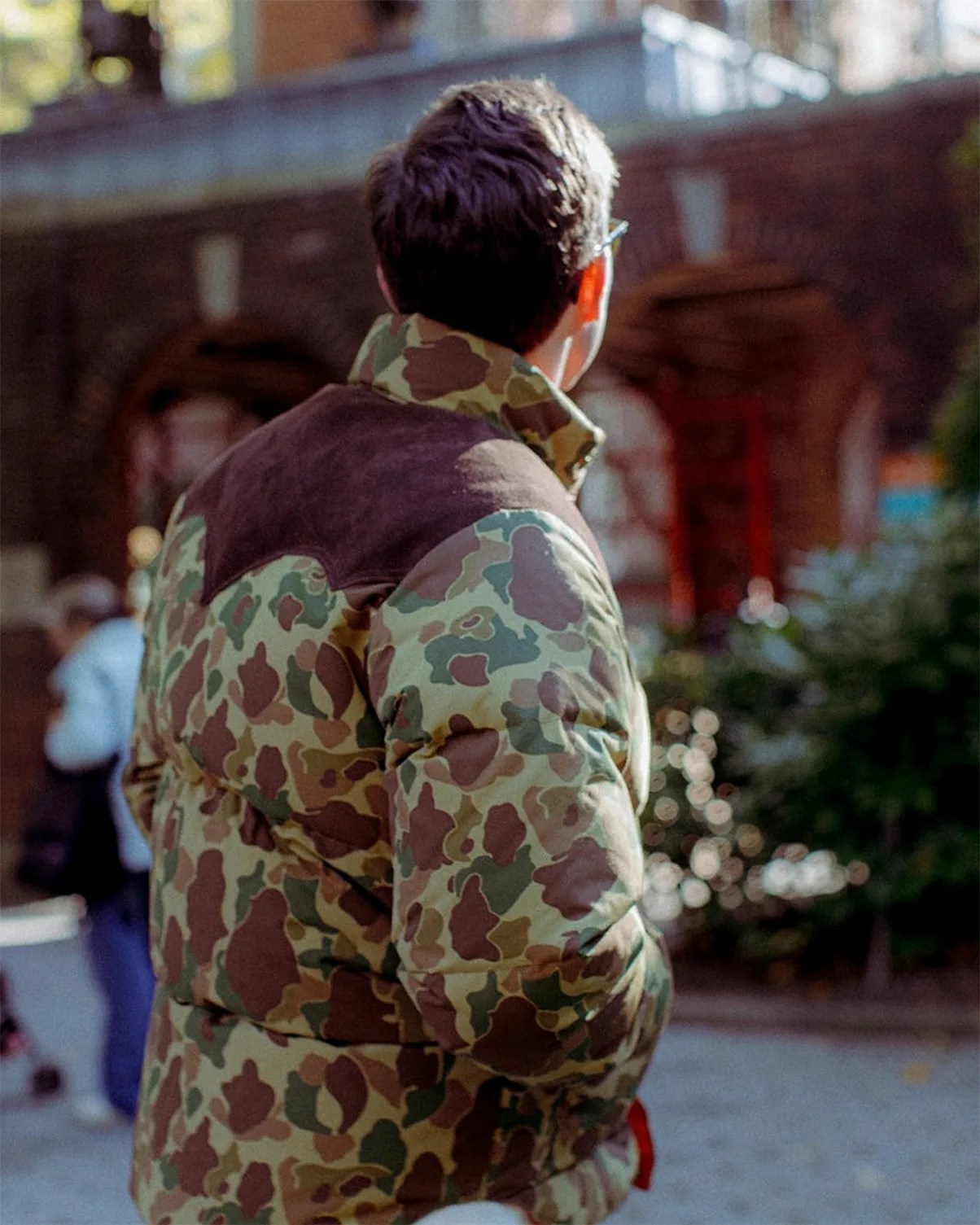 Person wearing a green and brown camouflage puffer jacket with a dark brown yoke, viewed from behind outdoors.