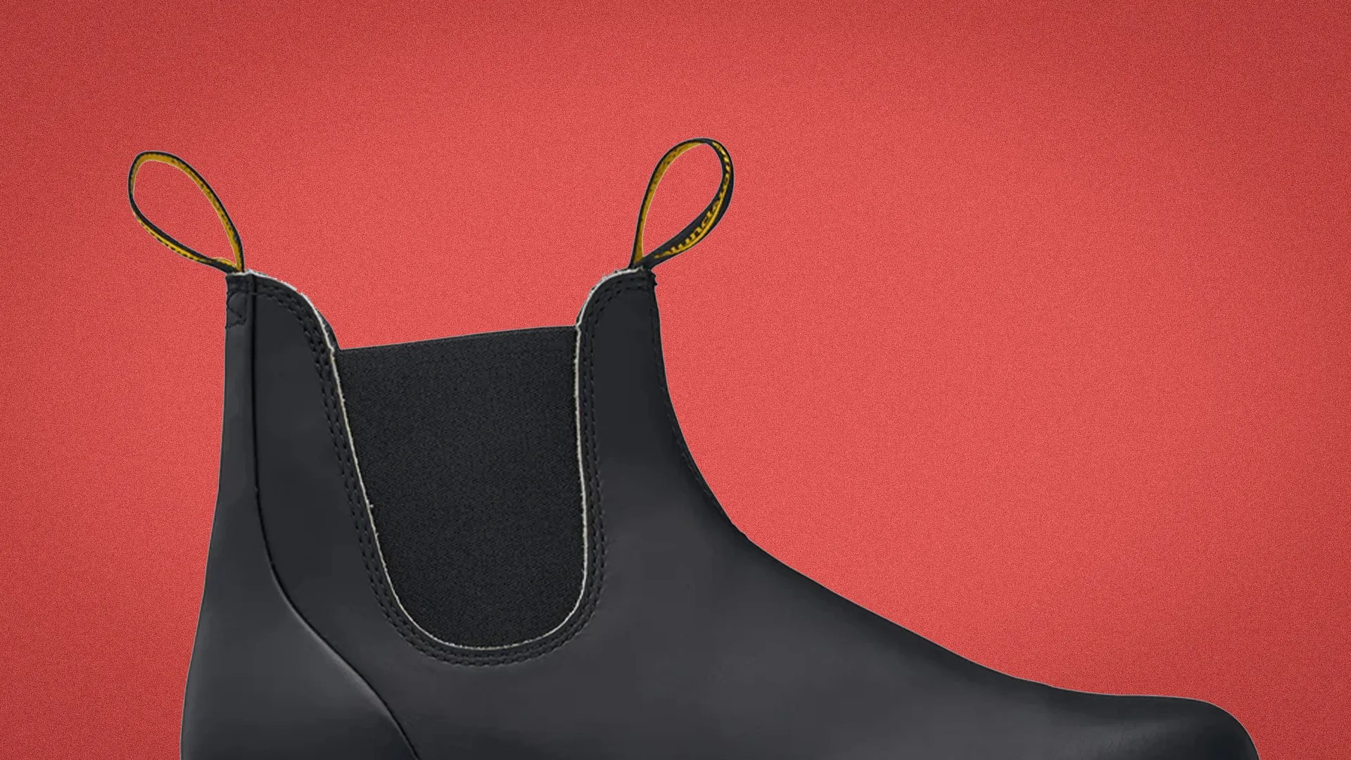 Black leather Chelsea boot with elastic side panel and yellow pull tabs on a red background.