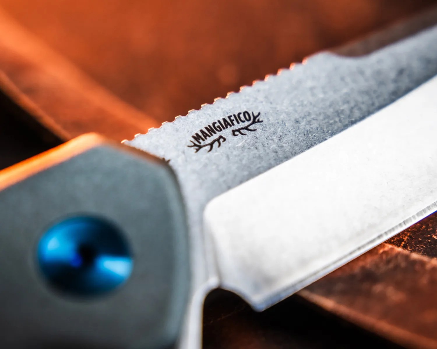 Close-up of a serrated knife blade with "Mangiafico" logo and a blue pivot screw on the handle.