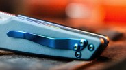 Close-up of a gray folding knife with a blue metal pocket clip resting on a textured surface.