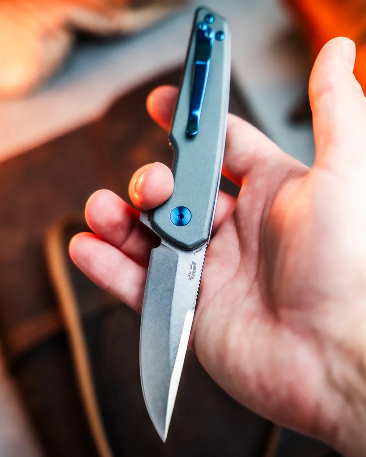 Hand holding a folding knife with a gray handle and blue clip and pivot screw.