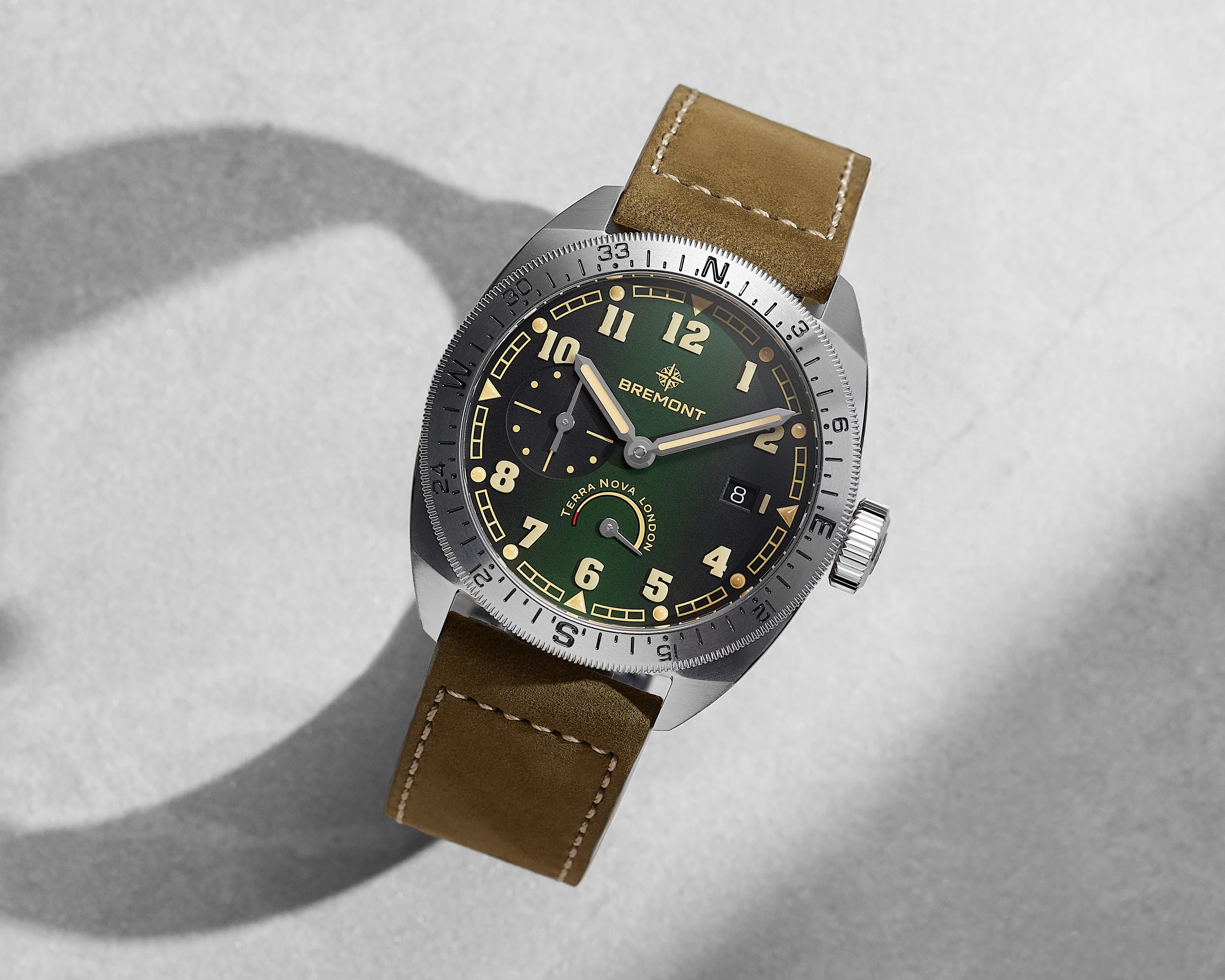 Green and black Bremont Terra Nova wristwatch with beige numerals, silver bezel, and brown leather strap on a gray surface.