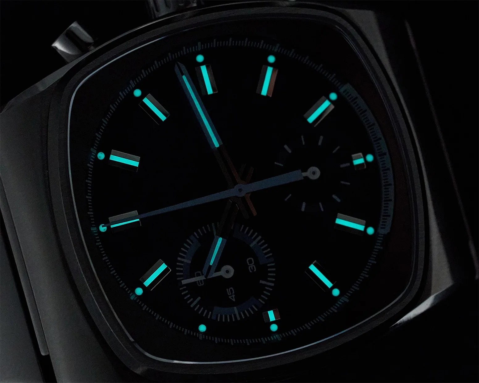 Close-up of a dark watch face with luminescent blue hour markers and hands glowing in low light.
