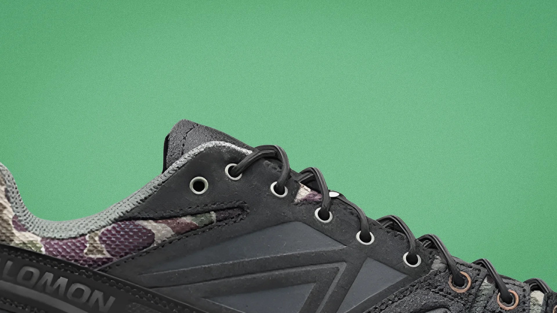 Close-up of a black and camouflage Salomon sneaker with black laces against a green background.