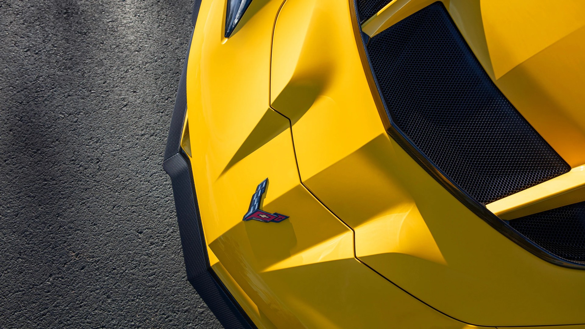 Close-up of a yellow sports car hood with a Corvette emblem and black mesh grille on asphalt.